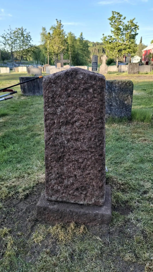 Slitt gravstein av stein på kirkegård, omgitt av gress og andre gravstøtter utendørs.